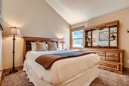 A cozy bedroom with a double bed, two bedside lamps, and a wooden dresser. The room has a beige color scheme and a window with a view of trees. A folded blanket is placed at the foot of the bed.