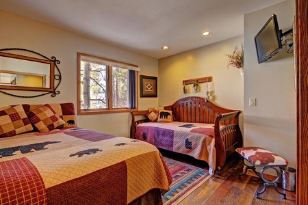 A cozy bedroom features two wooden beds with matching bear-themed quilts, a wall-mounted TV, a decorative mirror, a window with a view of trees, and wood flooring.