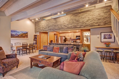 A cozy living room with two sofas, a coffee table, and an armchair. The room opens to a dining area and kitchen, featuring wood cabinets and modern appliances. Decor includes paintings and plants.