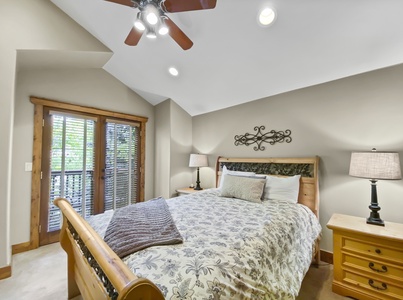 A bedroom with a wooden bed, two bedside tables with lamps, a ceiling fan, and double doors with blinds leading to a balcony. The room has light-colored walls and a vaulted ceiling.