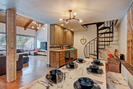 A modern open-plan dining and living area with a spiral staircase, wooden cabinetry, and a dining table set for six. A pink flamingo decor sits by the staircase, and a patio is visible in the background.