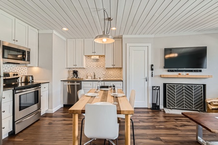 A modern kitchen and dining area with white cabinets, stainless steel appliances, wooden dining table, and four white chairs. A flat-screen TV and fireplace are visible in the background.