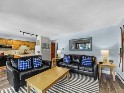 A living room with two black sofas, blue plaid pillows, a yellow coffee table, striped rug, and wall-mounted skis. The room opens to a kitchen with light wood cabinets and a black countertop.