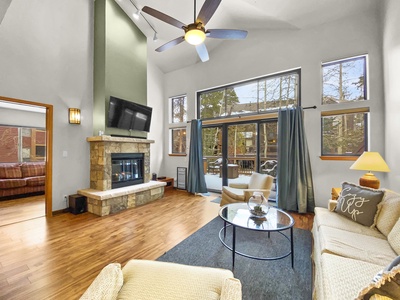 A spacious living room with high ceilings, a ceiling fan, large windows, a fireplace, and a wall-mounted TV. The room includes a beige sofa, armchair, coffee table, and sliding doors leading to an outdoor area.