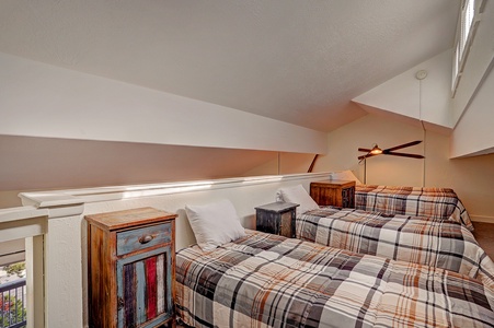 A loft bedroom with two single beds, plaid bedspreads, and two rustic nightstands under a sloped ceiling. A ceiling fan is visible above the beds.