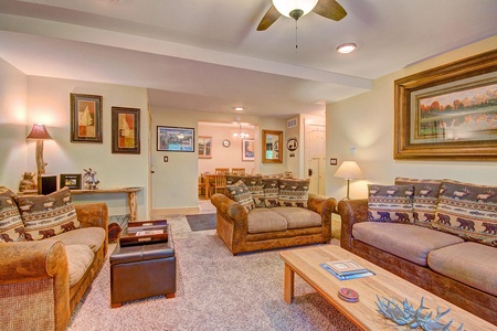 A cozy living room with two plush sofas, a coffee table, and wall art, leading to a dining area in the background. The room features warm lighting and a ceiling fan.