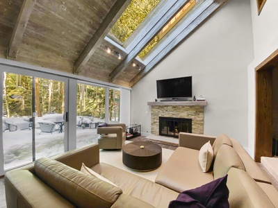 A modern living room with a vaulted wooden ceiling, a stone fireplace, a large sectional sofa, and large glass windows that provide a view of a snowy outdoors and trees.