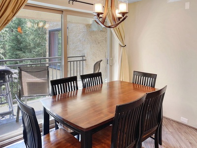 A dining room with a wooden table and six chairs is near a glass door leading to a balcony with outdoor furniture and a barbecue grill.