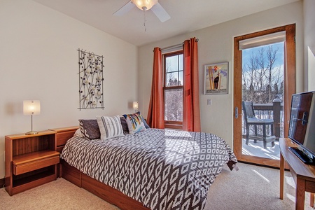 A cozy bedroom with a double bed, patterned bedspread, bedside tables with lamps, two windows with orange curtains, framed art, and a door leading to an outdoor deck with snowy landscape views.