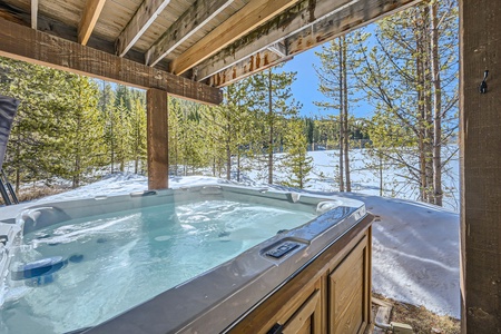 A hot tub sits under a wooden roof, overlooking a snowy landscape with evergreen trees and a partially frozen lake in the background on a sunny day.