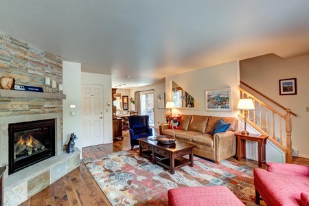 A cozy living room with a stone fireplace, a brown leather couch, a patterned rug, and wooden furniture. Stairs lead up from the right, and a dining area is visible in the background.