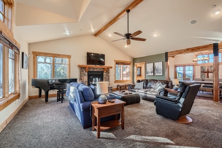 A spacious living room with a vaulted ceiling, furnished with sofas, recliner, a coffee table, and a grand piano. A ceiling fan and a fireplace with a TV above complete the cozy setup.