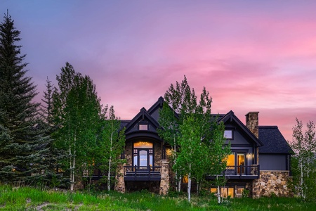 A two-story house with dark exterior colors and stone details is surrounded by trees at sunset, with a pink and purple sky in the background.