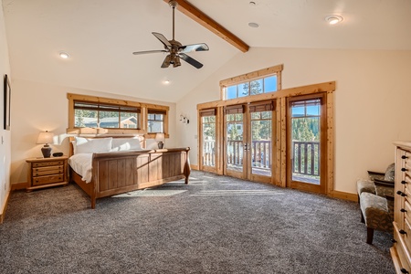 A spacious, carpeted bedroom with a wooden bed, nightstands, dresser, and bench. French doors and windows let in natural light and offer a view of an outdoor deck and forested area. Ceiling fan overhead.