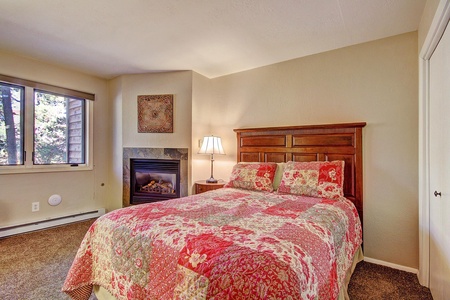 A bedroom with a wooden bed frame, floral quilt, bedside lamp, and a gas fireplace with a framed artwork above it. A window with a view of trees is on the left side of the room.