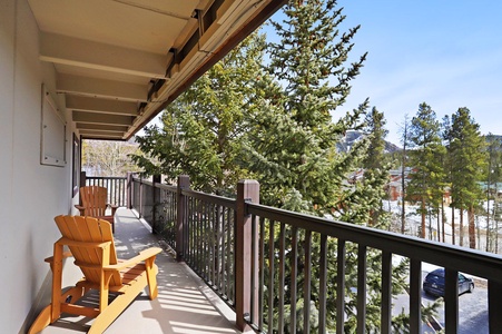 A balcony with two wooden Adirondack chairs overlooks a pine tree-filled landscape under a clear blue sky.