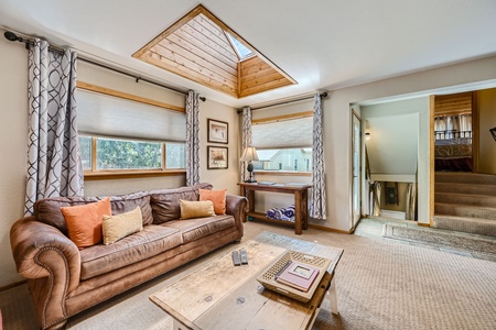 Living room with a brown sofa, assorted cushions, a wooden coffee table, and natural light from skylights and windows. Stairs in the rear lead to a different floor. Neutral decor with patterned curtains.