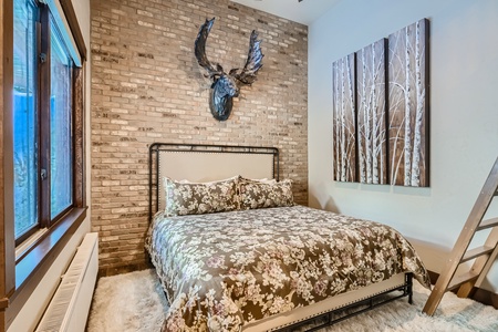 A bedroom with a floral-patterned bed, a brick accent wall, a moose head decoration, a large window, and a painting of birch trees. A ladder leans against the wall.