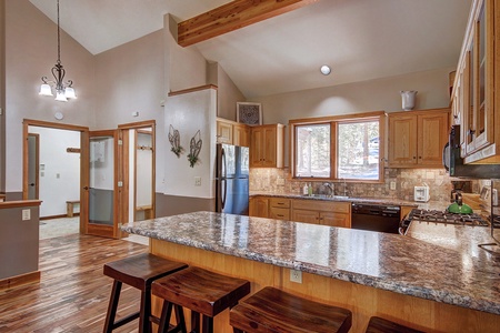 A modern kitchen with wooden cabinetry, granite countertops, and stainless steel appliances. The room features high ceilings, pendant lighting, and a view of trees through a large window.