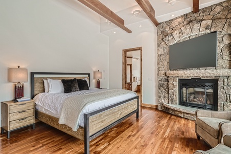 A cozy bedroom with a wooden bed, stone fireplace, TV, hardwood floors, two bedside tables with lamps, and a mix of modern and rustic elements.