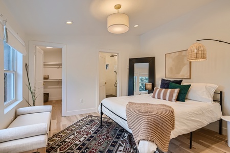 A modern bedroom with light walls, a bed with decorative pillows and a blanket, a large rug, an armchair, and a full-length mirror. The room includes an en-suite bathroom and walk-in closet.