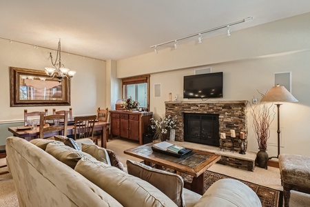 A cozy living room with a stone fireplace, wall-mounted TV, and beige couches. A wooden dining table with chairs is in the background, and a large mirror hangs on the wall beside a window.