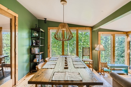 A dining room with green walls, a wooden table set with placemats, a modern chandelier, large windows, a bookshelf, and a floor lamp.