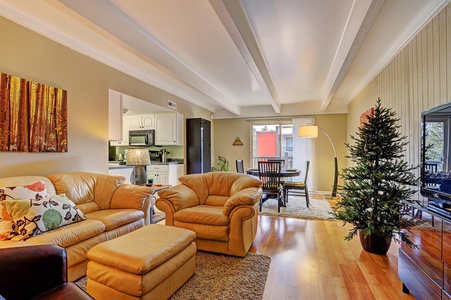 A living room with beige walls features a tan leather sofa set, a coffee table, a flat-screen TV, a small Christmas tree, and an adjacent dining area with a round table and chairs.