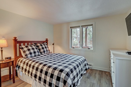 A small bedroom with a wooden bed frame and a black-and-white checkered bedspread, a bedside table with a lamp, a window, a white dresser, and a wall-mounted TV.