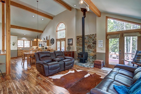 A cozy living room with leather seating, wooden flooring, a cowhide rug, large windows, a stone fireplace, and an adjoining dining area with a wooden table and chairs.