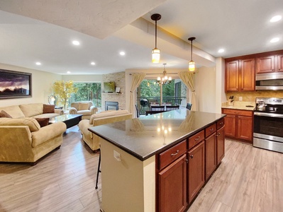 An open-concept kitchen with a large island, dark countertops, and wooden cabinets overlooks a well-lit living area with beige sofas and a fireplace, adjacent to large glass doors leading outside.