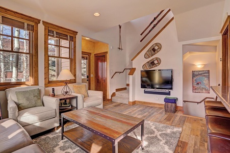 A cozy living room with wooden accents features a TV mounted on the wall, a wooden coffee table, two armchairs, a couch, and a staircase leading upstairs. Snowshoes and artwork adorn the walls.