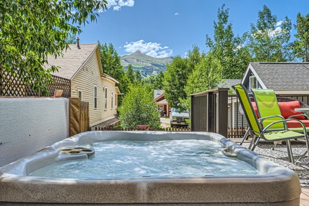 A hot tub sits on a wooden deck with trees and houses nearby, a mountain peak visible in the distance under a clear blue sky.