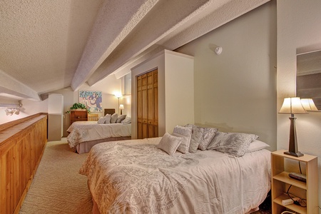 A cozy loft bedroom with two double beds adorned with gray bedding, wooden closets, and a bedside lamp. A dresser with plants and a framed picture are in the background.