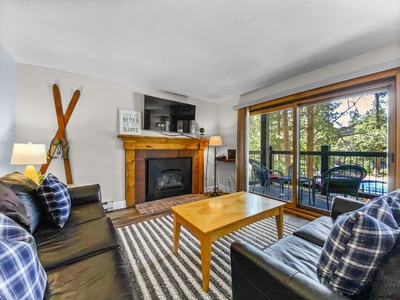 A cozy living room features a fireplace, a wooden coffee table, two black sofas, and a wall-mounted TV. Large windows and sliding glass doors provide a view of the outdoor balcony and wooded area.