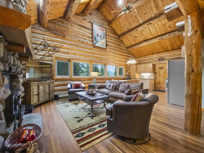 A cozy log cabin living room with a stone fireplace, leather sofas, wooden furniture, a rug, and high wooden ceilings. Natural light enters through multiple windows. Kitchen is visible in the background.