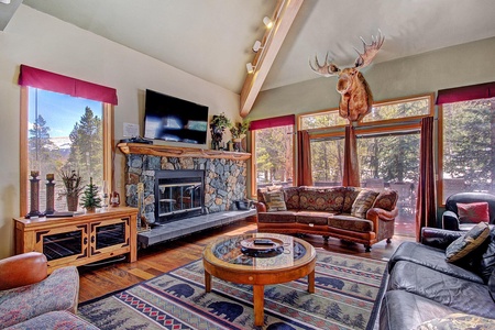 A cozy living room with a stone fireplace, mounted moose head, wooden furniture, and large, floor-to-ceiling windows covered with red curtains, offering a scenic outdoor view.