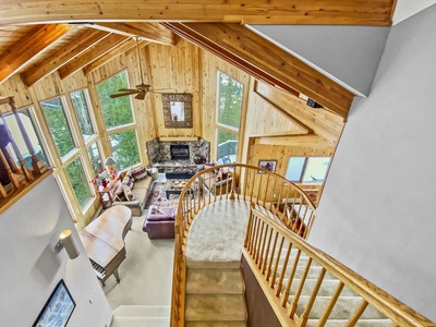 A bird's-eye view of a spacious log cabin living area with wooden beams, a stone fireplace, large windows, and a spiral staircase leading down to a cozy seating area.