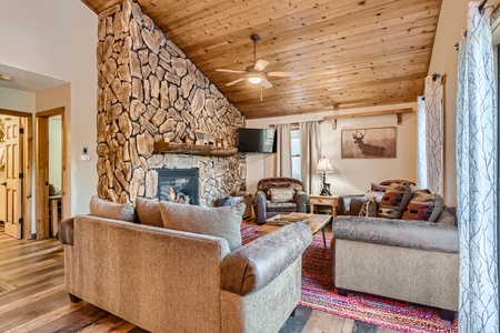 Cozy living room with stone fireplace, wooden ceiling, armchairs, sofas, a colorful rug, TV mounted on the wall, and natural light from windows with curtains. Artwork featuring a deer hangs on the wall.