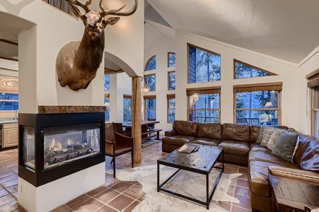 A cozy living area features a mounted deer head, a sectional sofa, a glass-encased fireplace, and large windows showcasing a forested view. A dining area and kitchen are visible in the background.