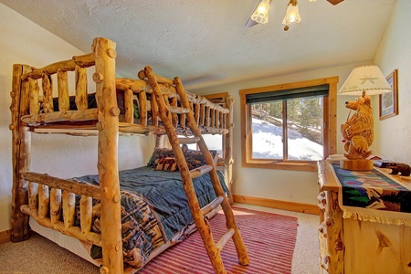 A rustic bedroom with wooden bunk beds, a window with a snowy outside view, a bear-themed lamp, and a striped red rug on the floor.