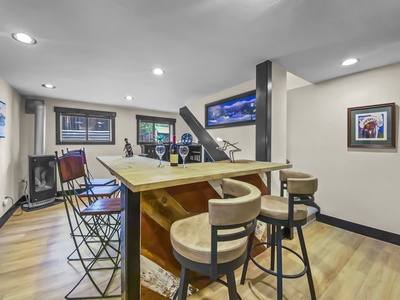 A modern basement bar area with wooden floors, a high table with bar stools, wall art, and seating next to windows.