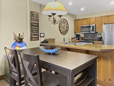 A kitchen with wooden cabinets, stainless steel appliances, and tiled countertops. The foreground shows a rectangular dining table with two chairs, a bowl of dried pinecones, and a wall clock above.