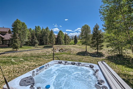 A bubbling hot tub on a patio overlooks a grassy yard surrounded by trees, with houses and a clear blue sky in the background.