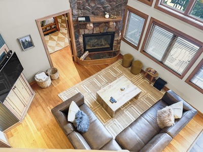 An overhead view of a cozy living room with a brown sofa, wooden coffee table, stone fireplace, and large windows. The room features wooden floors, a beige rug, and rustic decor.