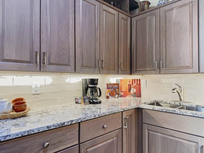 A kitchen with gray cabinets, white tile backsplash, and a granite countertop. There is a coffee maker, cups on a tray, a cookbook, and a sink.