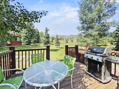 A wooden deck features a round glass table with green chairs and a gas grill. The deck overlooks a spacious, green lawn with trees and a clear blue sky.