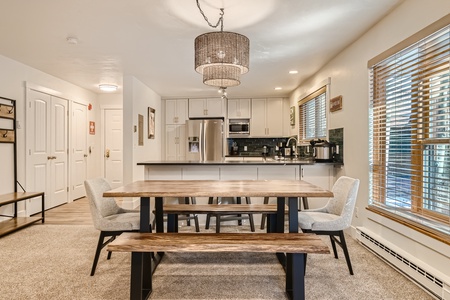 A modern kitchen and dining area featuring a wooden dining table with benches, a chandelier, stainless steel appliances, white cabinets, and a window with blinds.