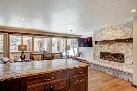 A modern living room features a stone fireplace, large windows with a snowy view, a wall-mounted TV, a leather sofa, and a kitchen island in the foreground.