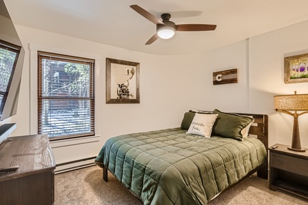 A small bedroom with a green bedspread, wooden headboard, side table with lamp, framed pictures on the walls, a ceiling fan, and a window with blinds.